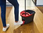 Person in jeans and white sneakers using a red and black mop bucket with a foot pedal.