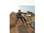 Homem sentado numa cadeira de campismo Crivit à mesa com comida junto ao mar.