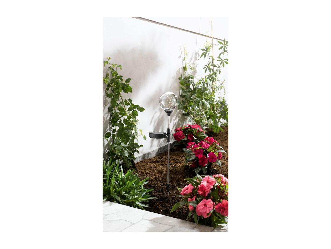 Solar-powered garden stake light with a glass globe, surrounded by pink flowers and green plants.