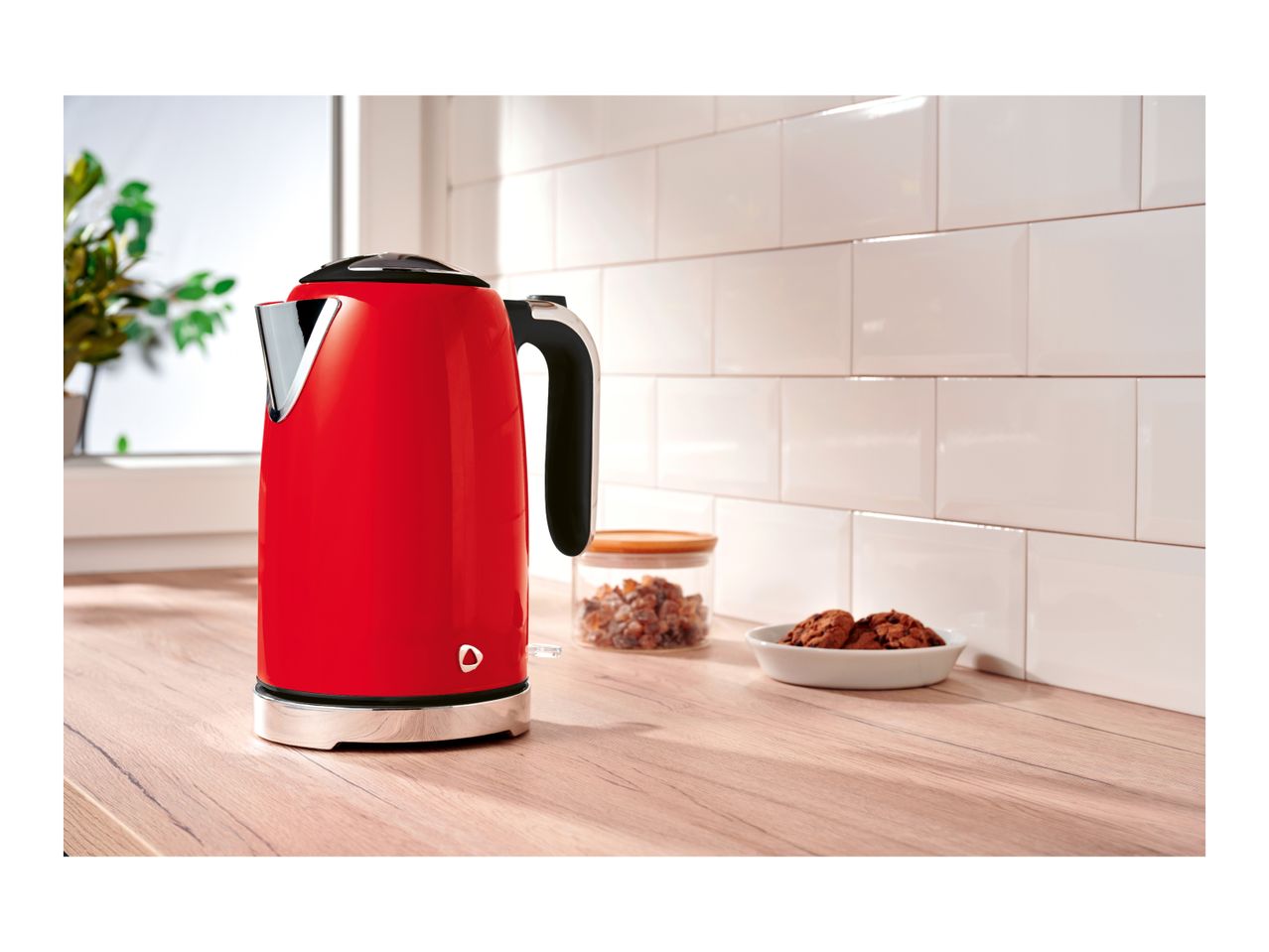 Red electric kettle on a wooden kitchen countertop next to cookies and a jar.
