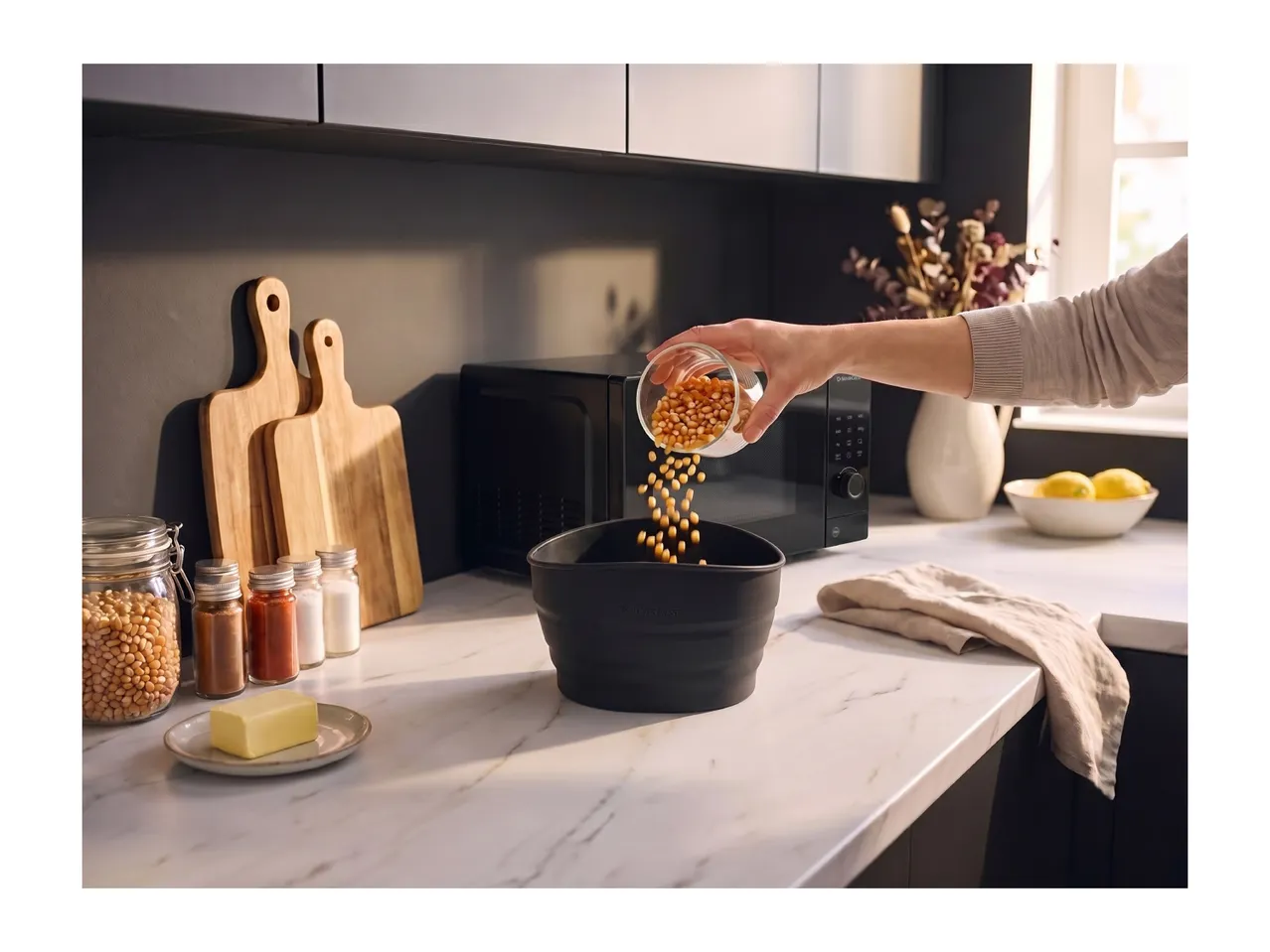 Person pouring popcorn kernels into a black bowl on a kitchen counter with a microwave.