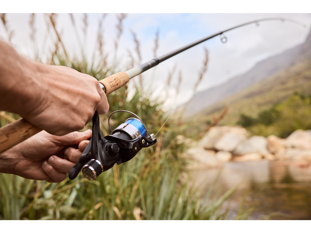 Καλάμι και μηχανισμός ψαρέματος CRIVIT με μπλε πετονιά, κρατημένα από χέρια, κοντά σε ποτάμι.