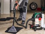A person using a wet/dry vacuum cleaner to clean a car mat in a garage.