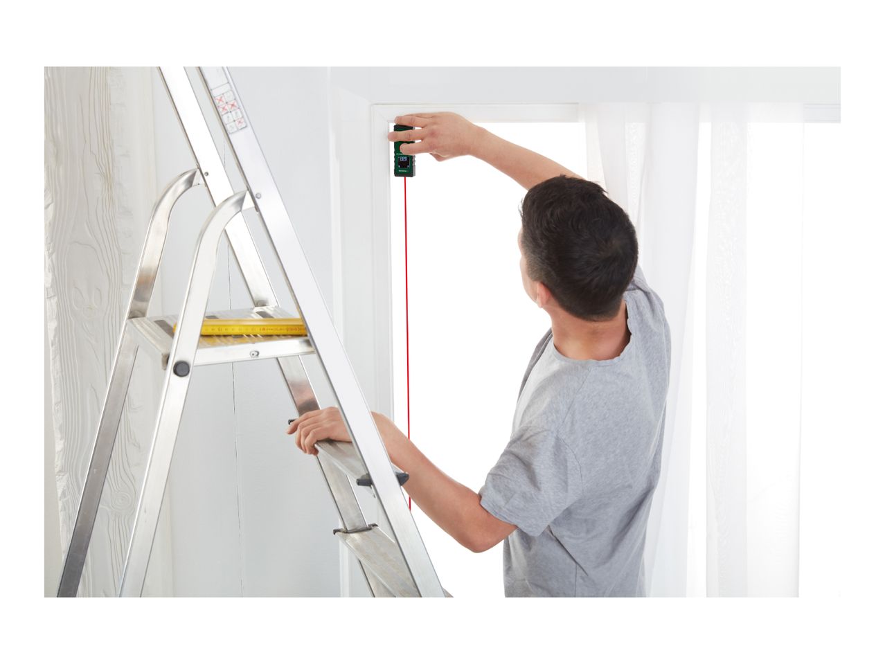 Man on a ladder using a laser distance meter to measure a window frame.