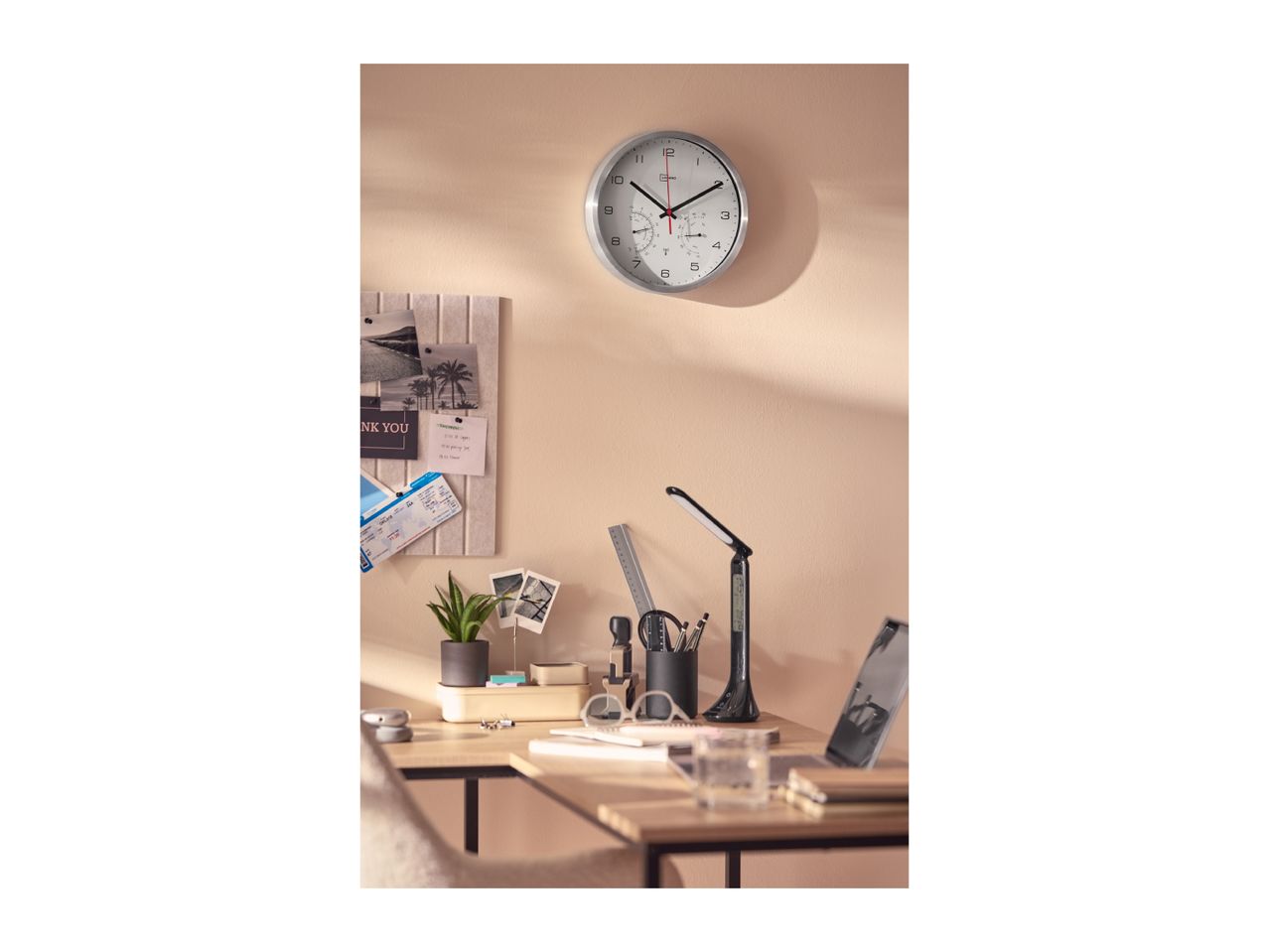 Wall clock with thermometer and hygrometer above a desk with office supplies.