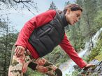 Woman in a black puffer vest and red fleece top, climbing a rock near a waterfall.