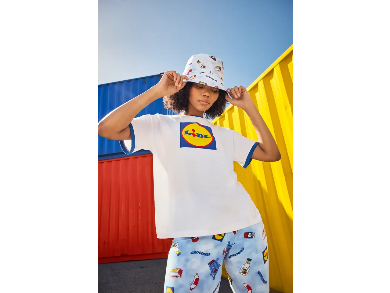 Woman in a white t-shirt with a large logo, bucket hat, and patterned shorts with groceries.