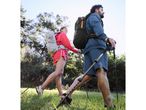 Two hikers with backpacks and trekking poles walk through a grassy, wooded area.