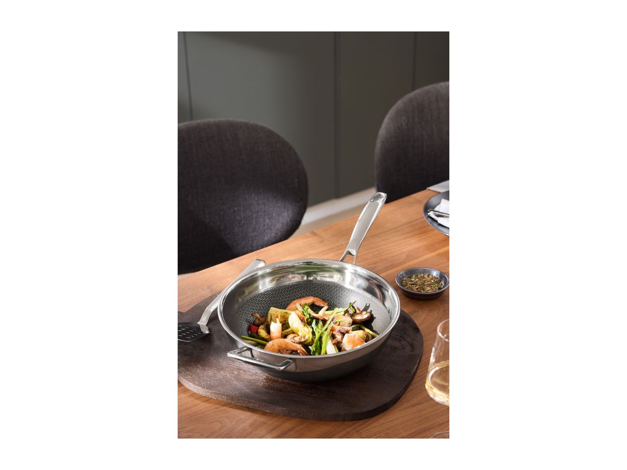 Stainless steel wok with stir-fry, spatula, and spices on a wooden table.