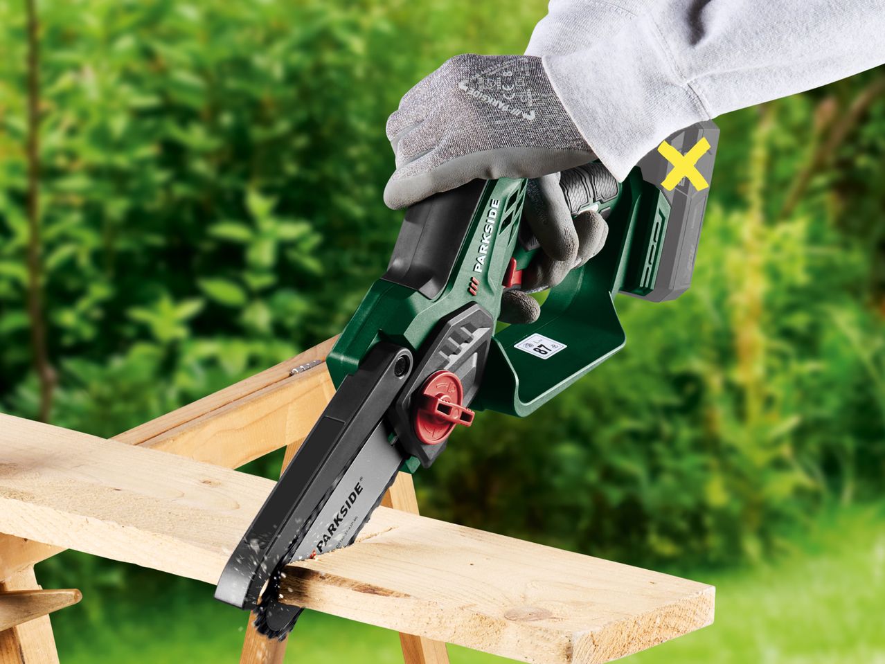A person wearing gloves uses a cordless mini chainsaw to cut a wooden plank outdoors.