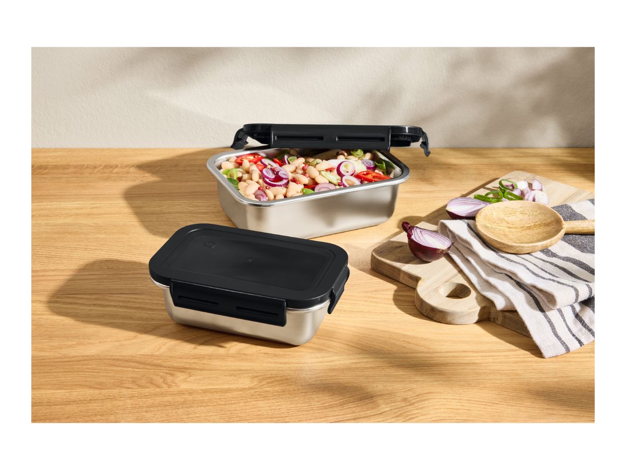 Two stainless steel food containers, one open with a bean salad, on a wooden table with chopped onions.