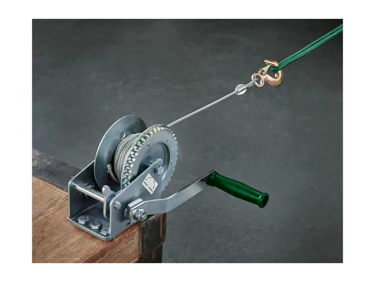 A hand winch with a green handle, steel cable, and hook, mounted on wood.