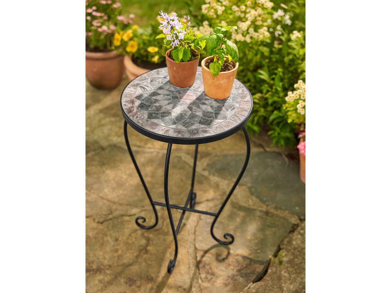 A mosaic-top outdoor side table with two potted plants on a stone patio.