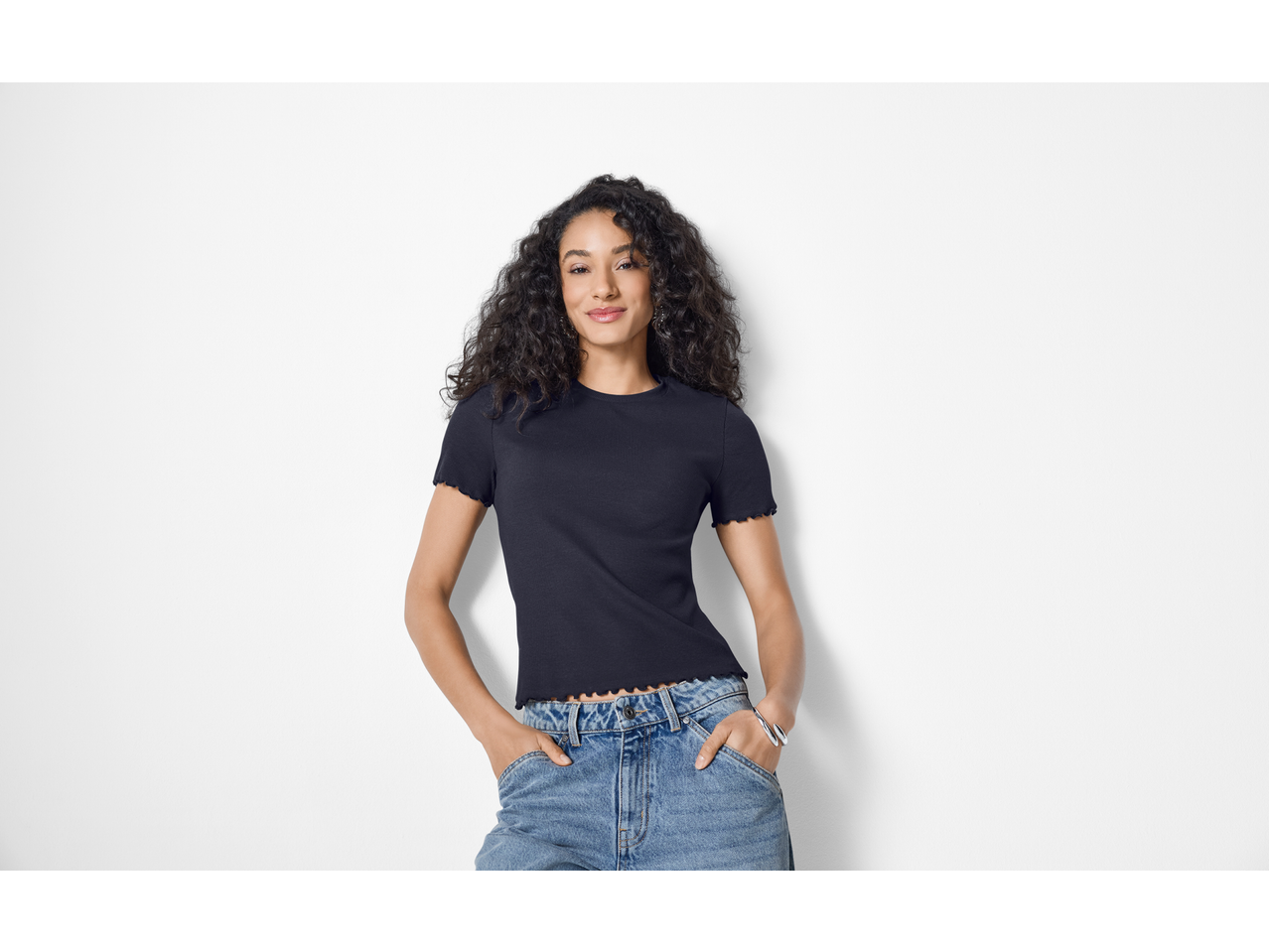 Femeie zâmbitoare, cu păr creț, purtând un tricou reiat ESMARA negru și blugi.