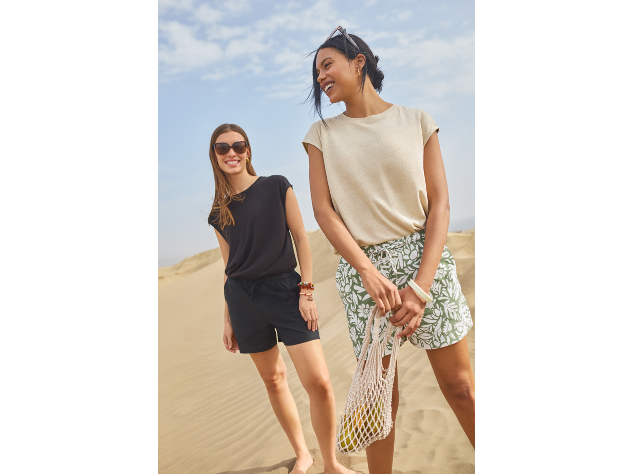 Two women in casual summer tops and shorts, one holding a net bag with fruit, on a sandy beach.