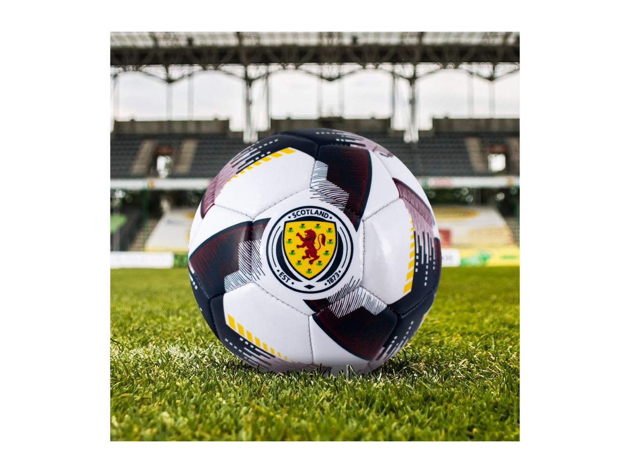 A football with a Scottish crest on a green field in a stadium.