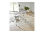 A white patterned ironing board standing in a bright room with a laundry basket.