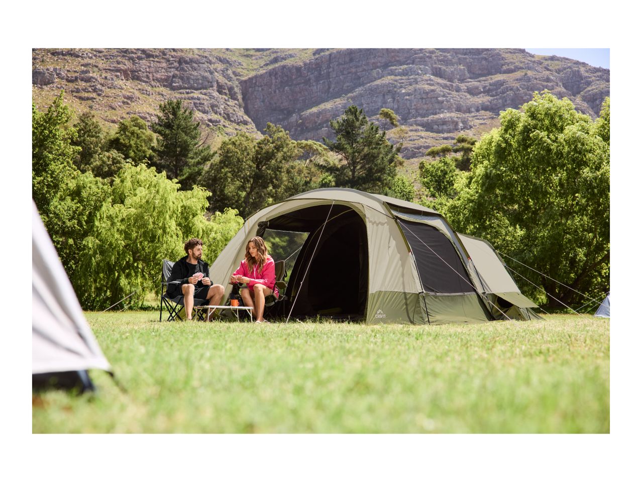 Two people playing cards outside a large camping tent in a grassy area with mountains.