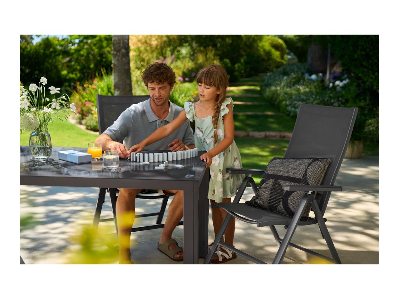 Father and daughter playing dominoes on a garden table with chairs in a sunny garden.