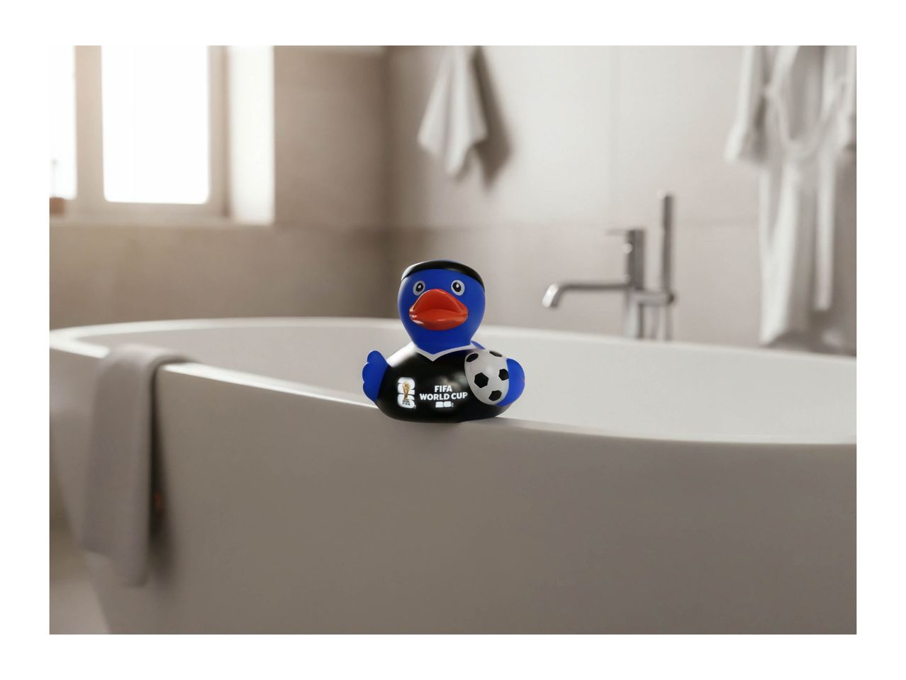 A blue rubber duck wearing a black shirt with 'FIFA World Cup' text, holding a soccer ball, sitting on the edge of a bathtub.