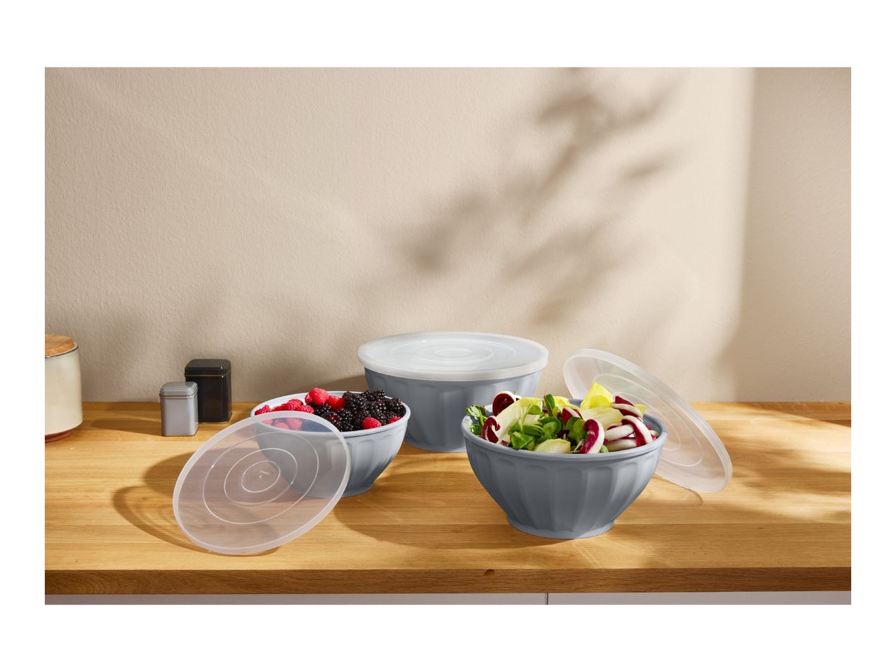 Three grey bowls with lids, two filled with berries and salad, on a wooden counter.