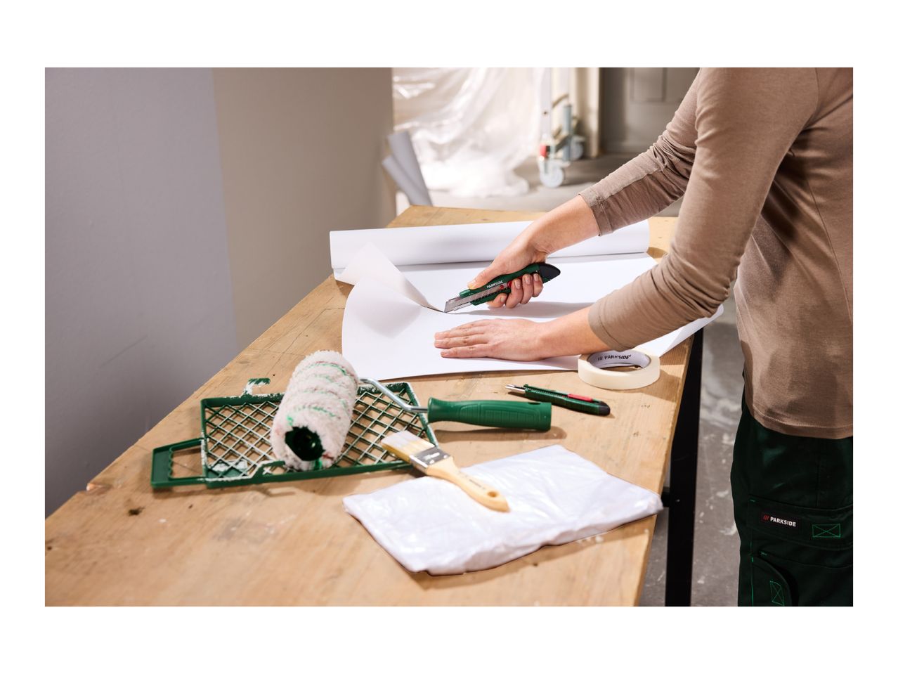 Person cutting wallpaper with a utility knife, with painting tools on a wooden table.