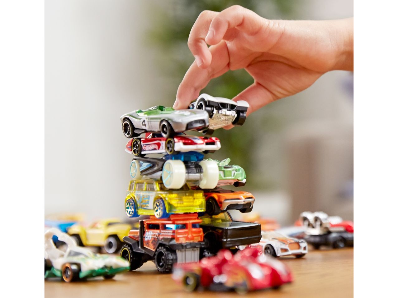 A child's hand places a Hot Wheels car on top of a stack of other toy cars.
