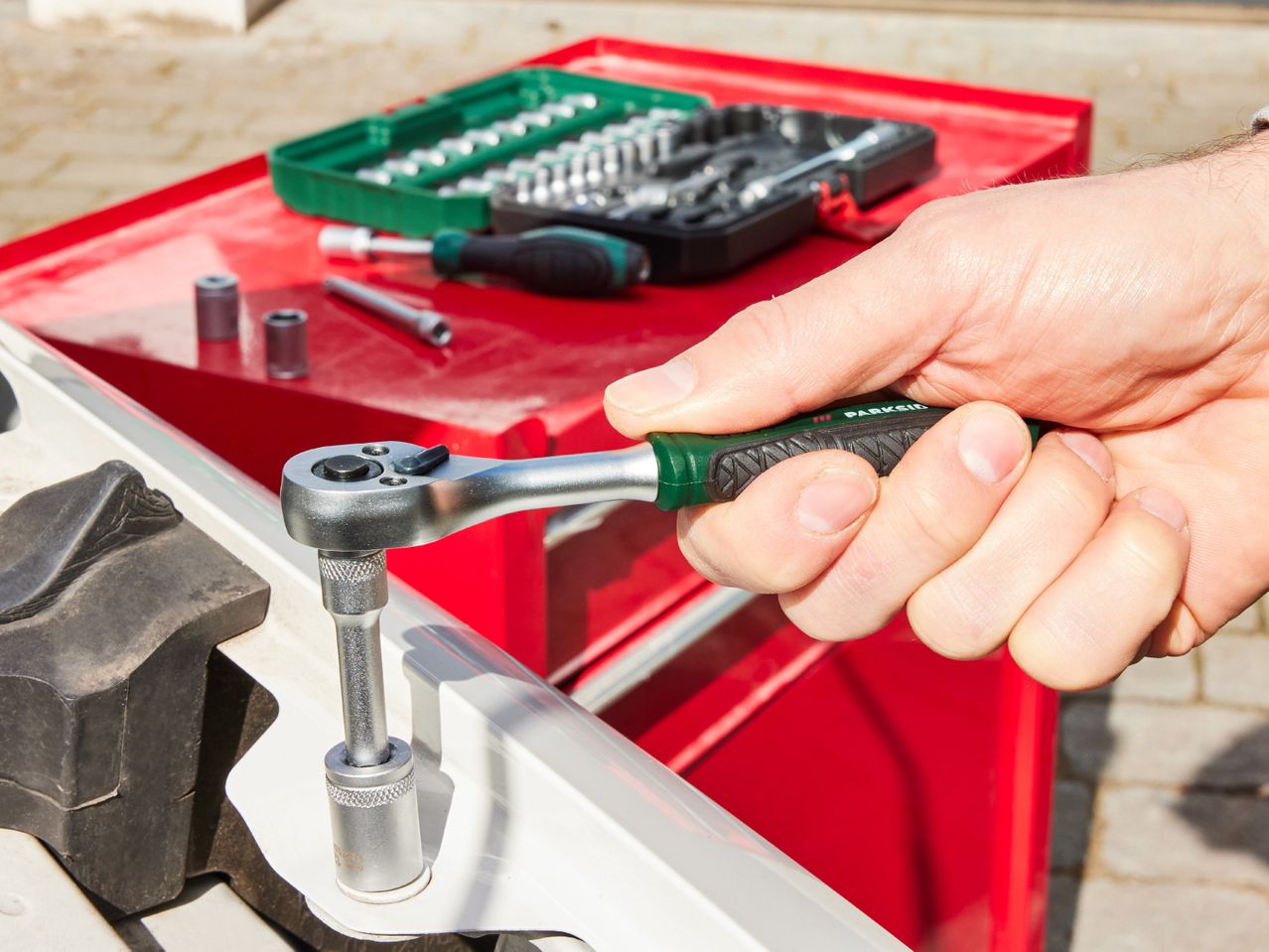 A hand uses a ratchet wrench on a metal part near a red toolbox filled with tools.