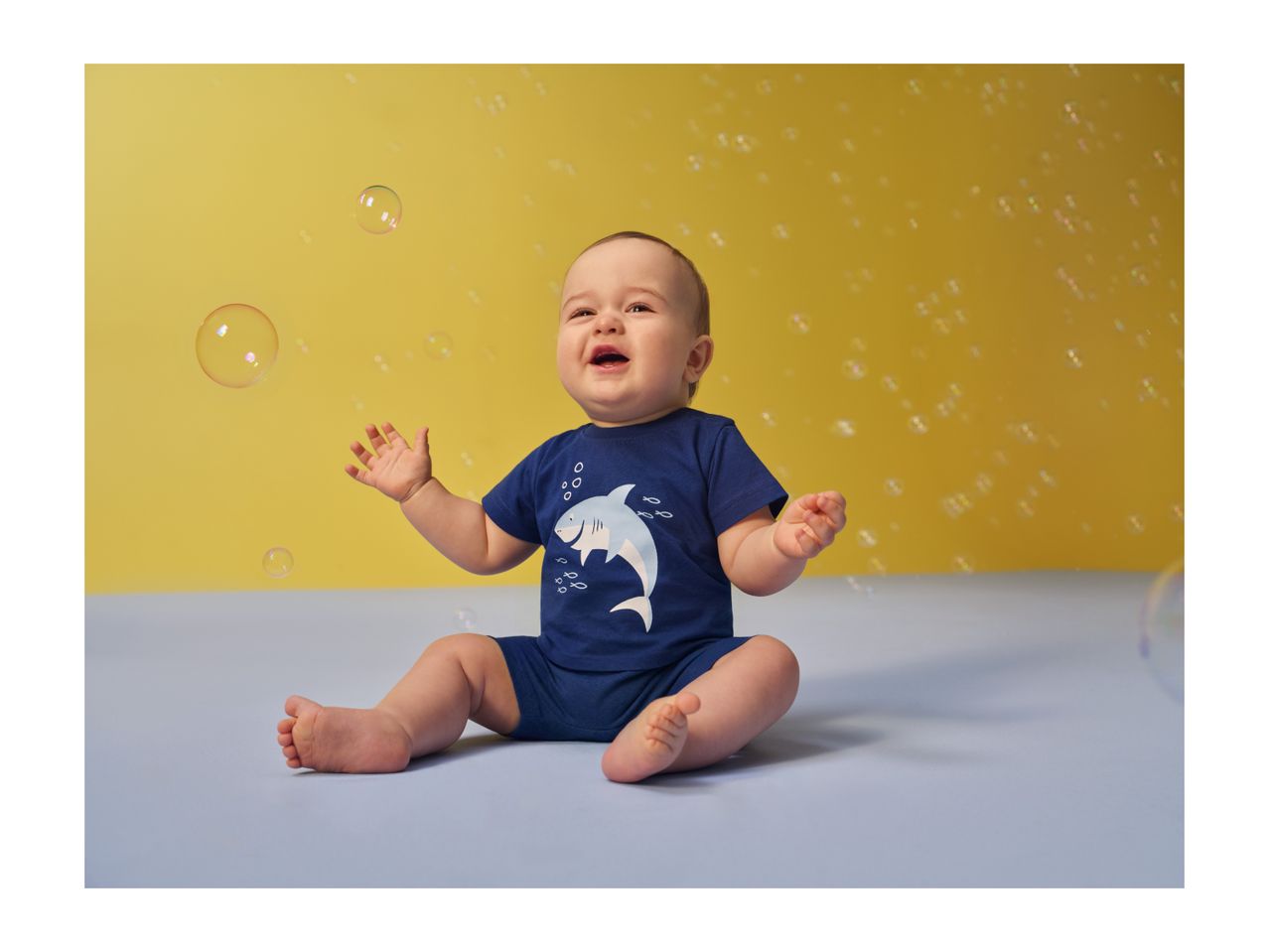 Happy baby in a blue shark-themed outfit, sitting on a light blue surface with bubbles.