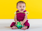 Ein glückliches Baby in weinroten Stramplern spielt mit einem grünen Spielzeug.