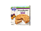 Alpenfest Plum Crumble Cake, 1250g, frozen, with a slice cut out, on a white wooden background.