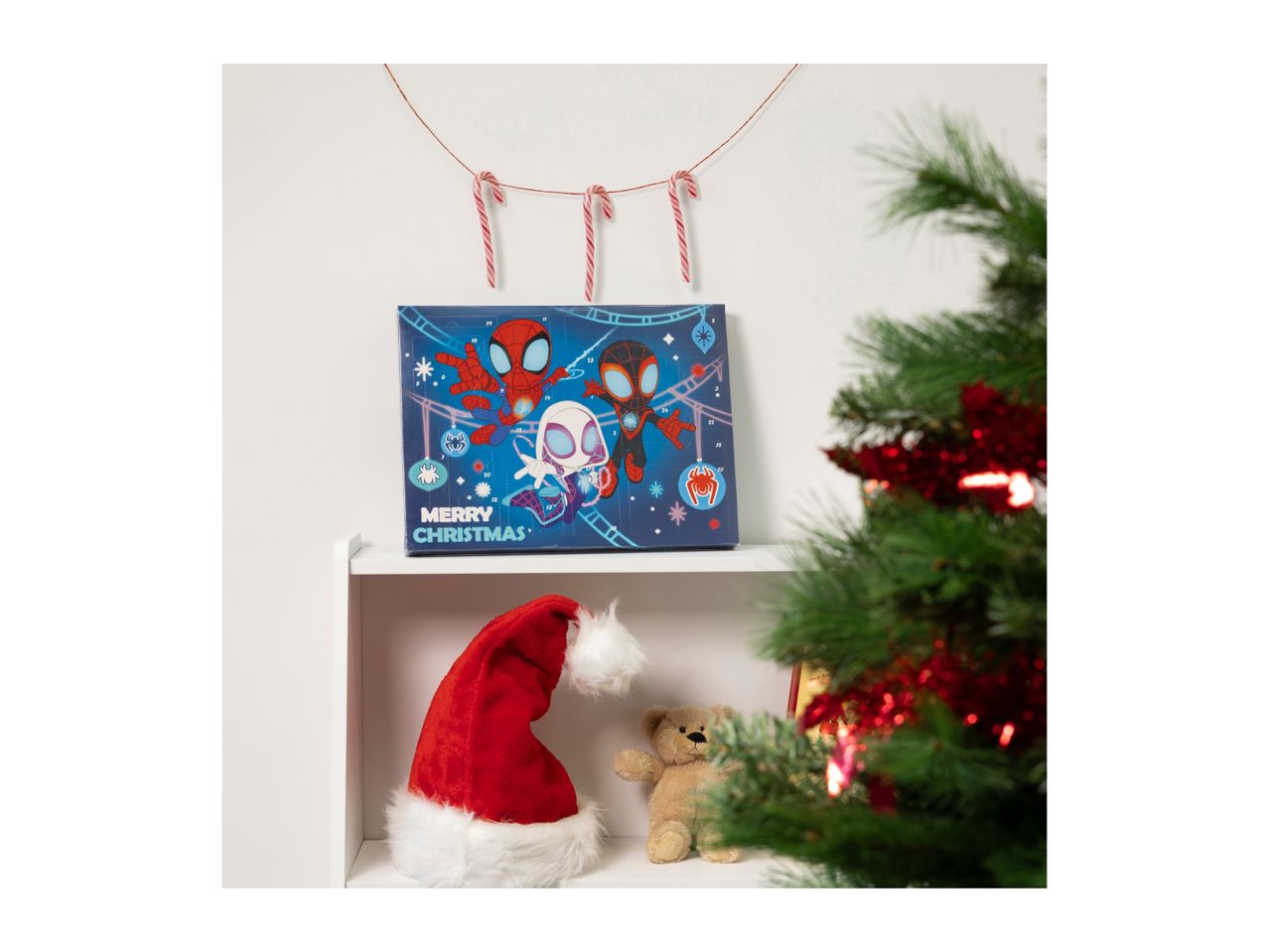 Spider-Man advent calendar, Santa hat, and teddy bear on a shelf with a Christmas tree.