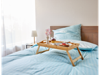 A bamboo breakfast tray sits on a bed with a light blue striped duvet, holding a dessert, toast, and flowers.