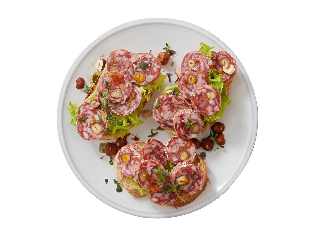Szalámis és mogyorós bruschetta salátával és mézzel egy fehér tányéron.