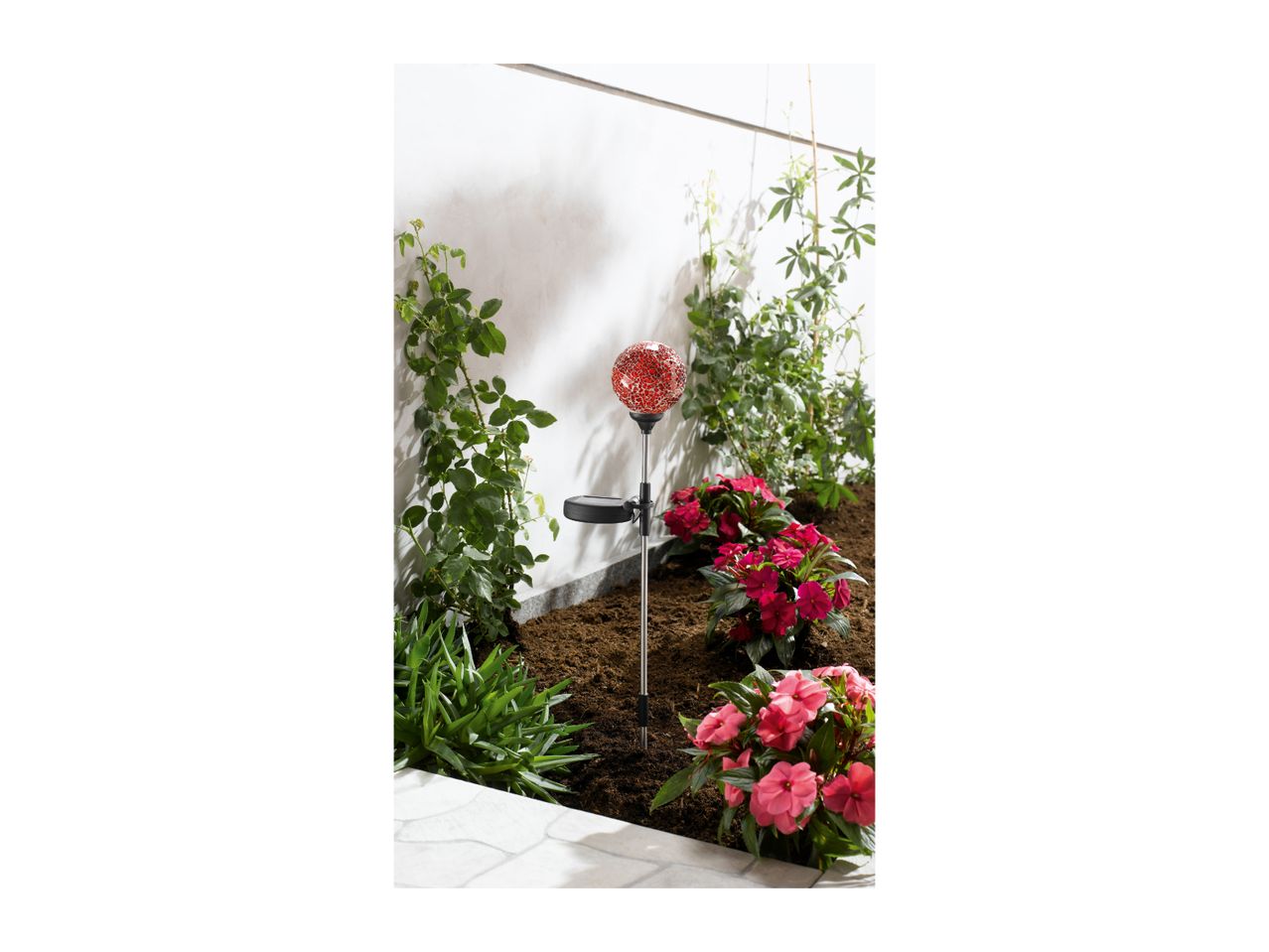 Solar garden light with a red mosaic globe in a garden bed with pink flowers.