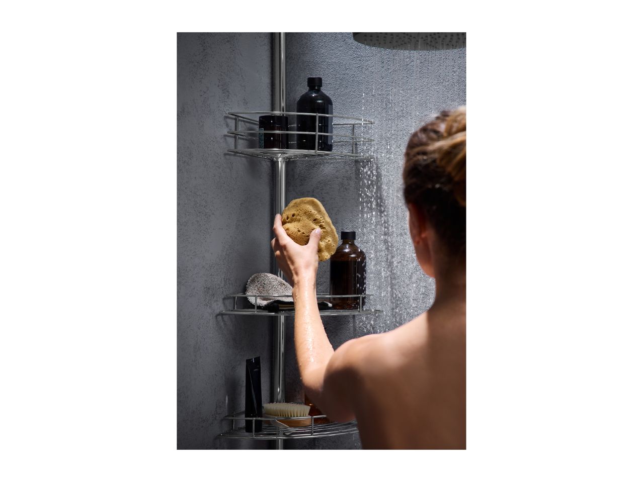 A person showering, reaching for a natural sponge on a shower caddy with various bath products.