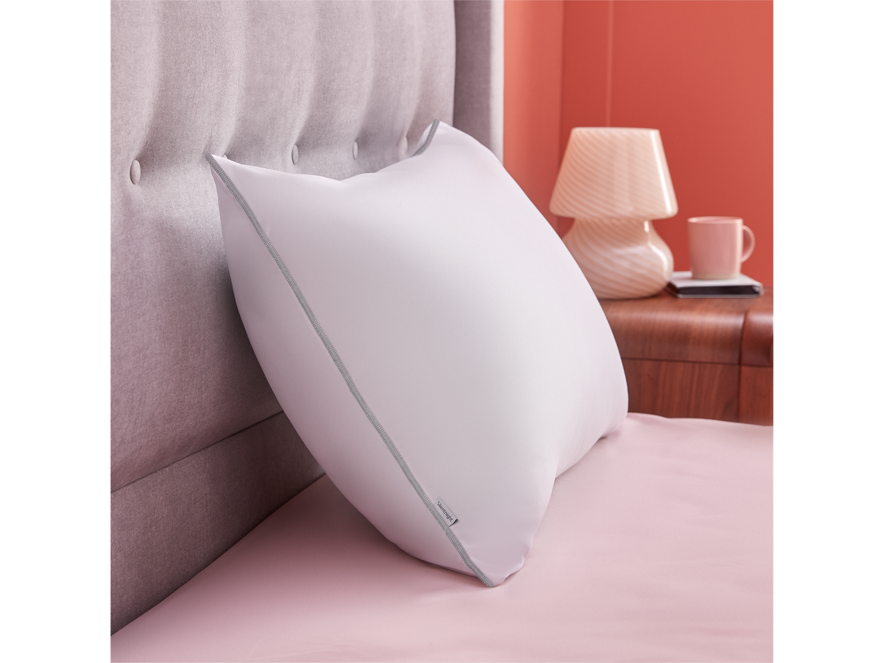 A Silentnight pillow sits on a blush pink bed, next to a grey headboard and a bedside table.