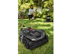 A robotic lawnmower on green grass, with a woman in the background using a smartphone.