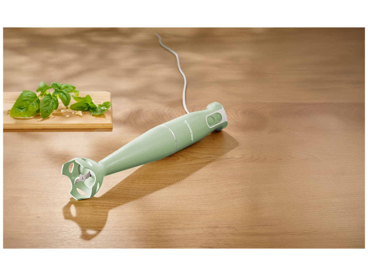 Mixeur plongeant vert sur une table en bois avec du basilic et du parmesan.