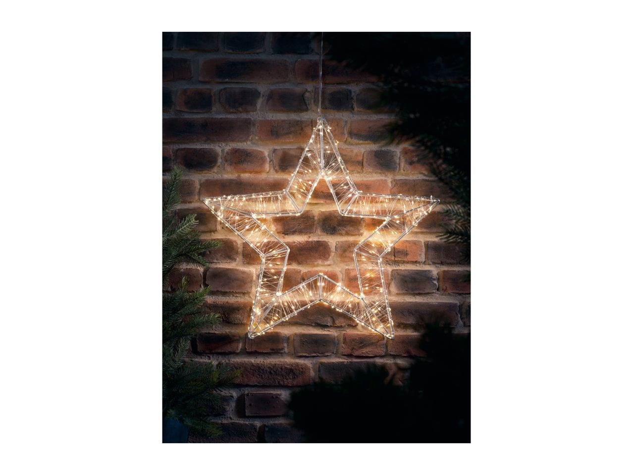 Illuminated star decoration hanging against a brick wall with pine branches