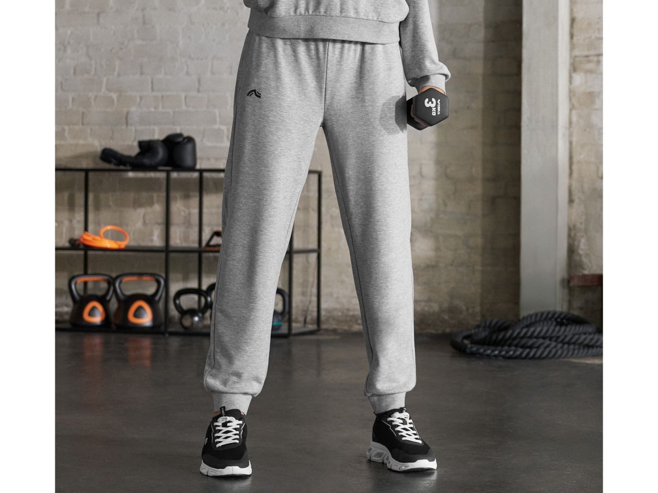 A person in gray sweatpants and sneakers holding a 3kg dumbbell in a gym setting.