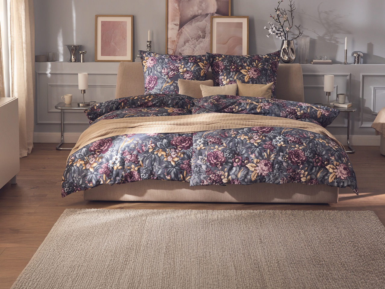 Спалня с флорално спално бельо в лилави и сиви тонове, с бежово одеяло и килим.