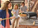 Tre amici preparano una pizza con un forno per pizza da esterno, su un tavolo di mattoni.