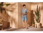 Homme en chemise grise à manches courtes et short en jean bleu clair, debout dans un coin ensoleillé avec un cactus.