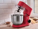 A red stand mixer with a whisk attachment and stainless steel bowl on a wooden counter with almonds and eggs.