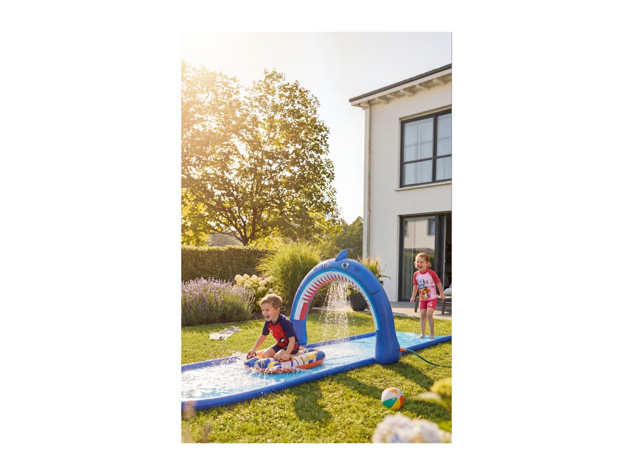 Two children playing on a shark-themed water slide in a sunny garden.