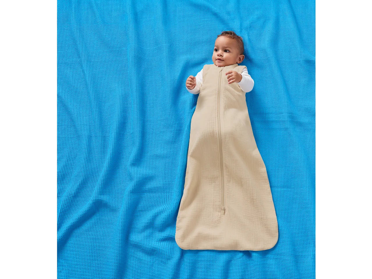 Bebeluș în sac de dormit din muselină LUPILU pe o pătură albastră.