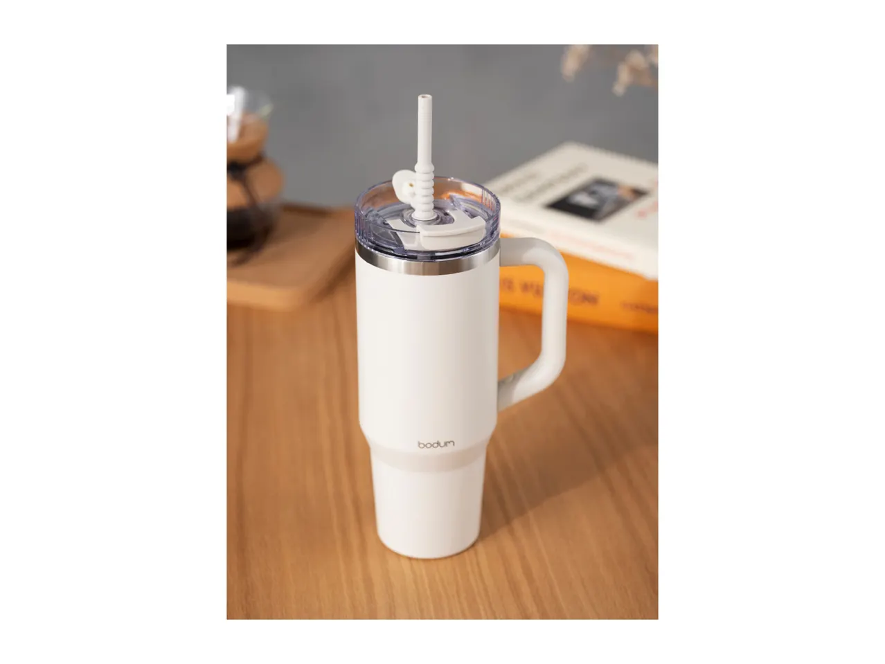 White travel mug with a handle and straw on a wooden table with a book in the background.