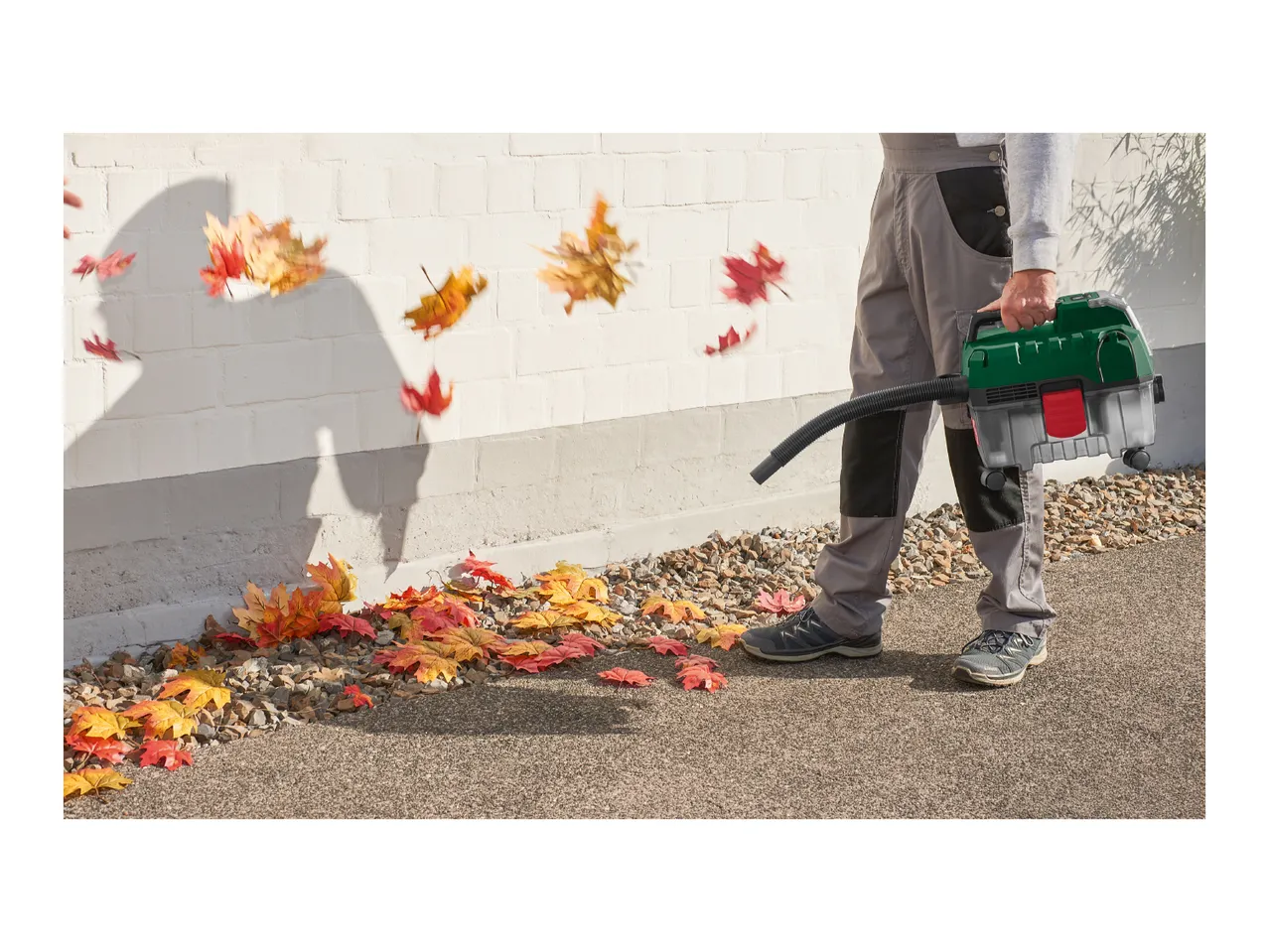 Person using a leaf blower to clear autumn leaves from a paved area next to a white wall.