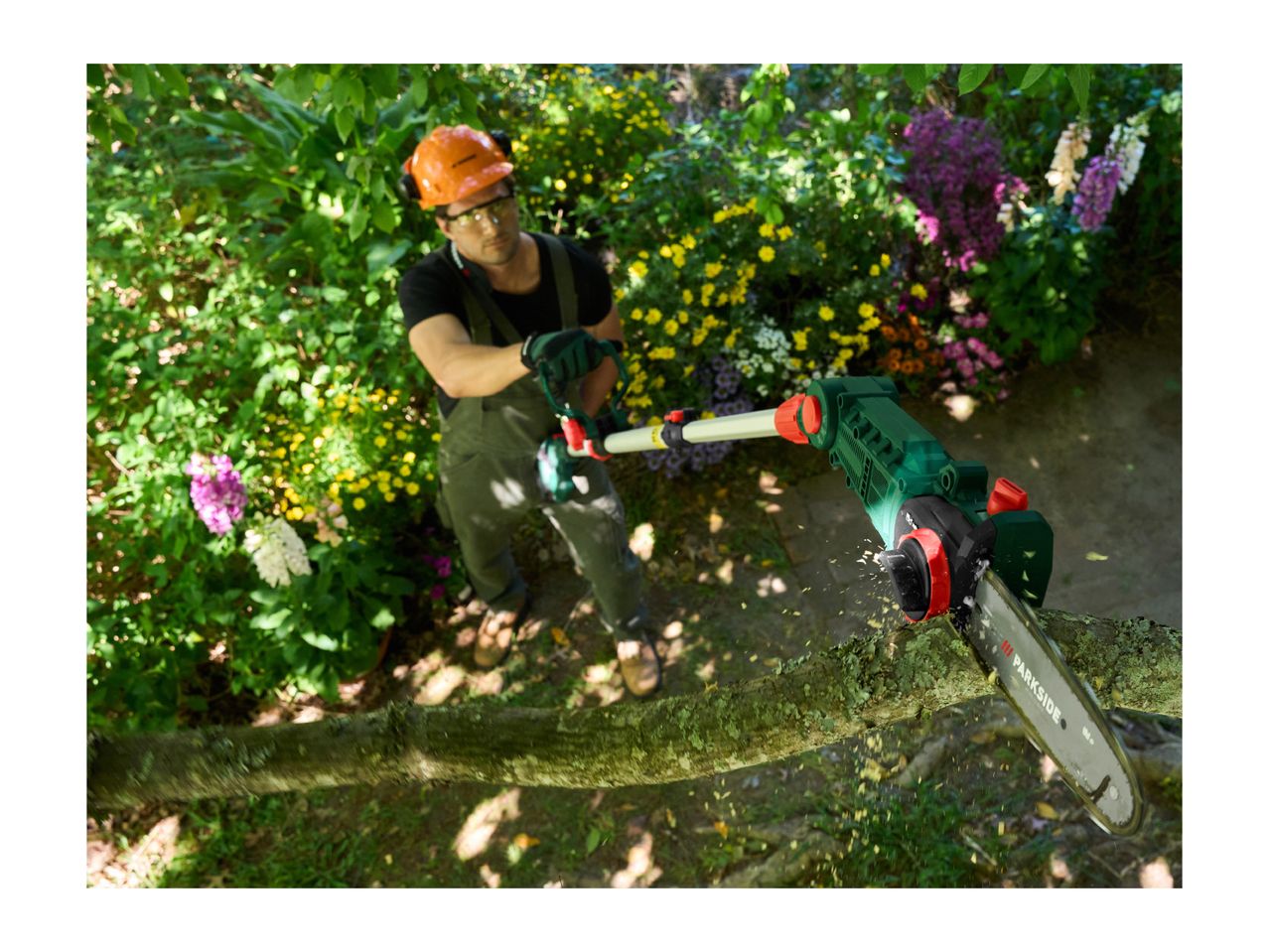 Man in protective gear using a pole chainsaw to cut a tree branch, with wood chips flying.